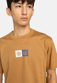 Camiseta de algodón marrón con cuello redondo, que presenta un diseño impreso en blanco y azul en la parte delantera con símbolos y formas geométricas.