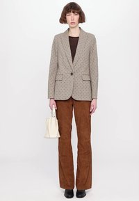 Lysbrun skræddersyet blazer med sorte polkaprikker, brune ruskindsbukser og en hvid håndtaske. Sorte sko fuldender outfittet.