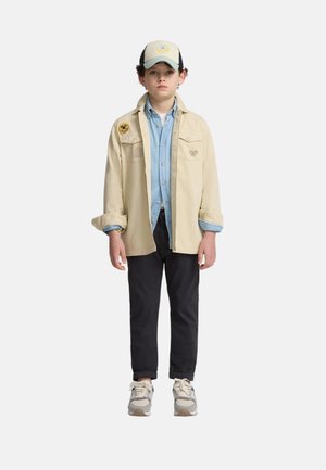 Chaqueta de camisa beige con bordado, camisa de mezclilla azul clara, pantalones negros, zapatillas grises y una gorra bicolor con un logo.