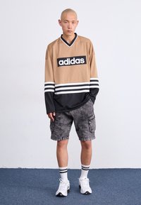 Νεαρός φαλακρός άνδρας που φοράει μαυροένδυτο μπλουζάκι με μακριά μανίκια από την Adidas, γκρι cargo σορτς, λευκές κάλτσες με ρίγες και λευκά αθλητικά παπούτσια που στέκεται σε εσωτερικό χώρο.