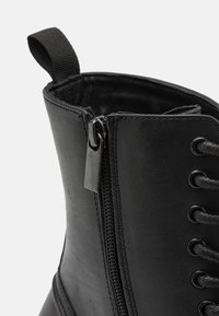 Botte de cheville en cuir noir avec une fermeture éclaire sur le côté, des lacets et une tirette en tissu ; texture lisse avec des détails de couture contrastés.