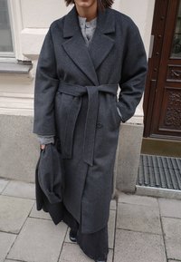 Pilkas vilnos mišinys paltas su diržu, plačiais atlapais ir dviem priekinėmis kišenėmis, uždėtas ant pilkos megztos viršūnės ir juodų džinsų sijono.