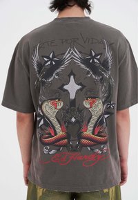 Grå oversized t-shirt med detaljeret grafisk design af slanger, fugle og et kors i livlige farver. "Ed Hardy" logo nederst.