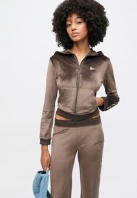 Survêtement en velours marron avec une veste zippée et un pantalon assorti. Comprend une capuche, des accents bronze et un logo sur la poitrine et la cuisse.