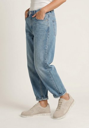 Osoba v světlých modrých riflech s ohrnutými nohavicemi a béžových teniskách se šňůrkami, stojící s rukama v kapsách před jednoduchým pozadím.