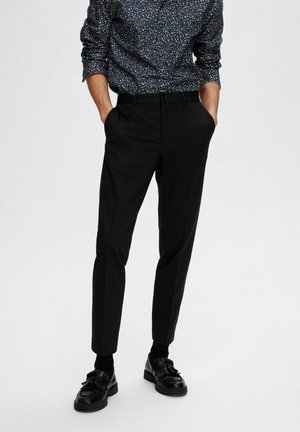 Pantalon noir ajusté avec une coupe droite et poches latérales, associé à des chaussures noires et une chemise à boutons bleu marine à motifs.