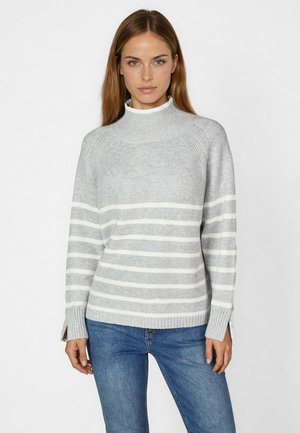 Grauer gestreifter Pullover mit hohem Kragen, versehen mit weißen horizontalen Streifen, gerippten Bündchen und seitlichen Schlitzen, kombiniert mit blauen Jeans.