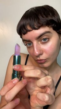 Lila glittrigt läppstift med spetsig topp, förpackat i ett glänsande grönt hölje. Höljet har en vridmekanism för applicering.