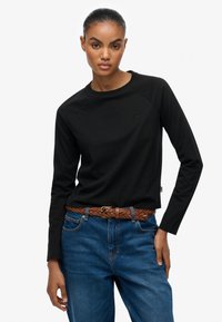 Zwarte longsleeve T-shirt met ronde halslijn en raglanmouwen, gecombineerd met een blauwe jeans en een bruin gevlochten riem. Subtiele branding op het shirt.