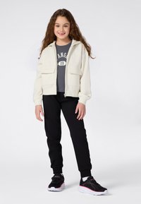 Ragazzina in piedi, che indossa una giacca bianca con zip, una maglietta grigia con una grafica, pantaloni neri e sneakers nere con suola bianca e dettagli rosa.