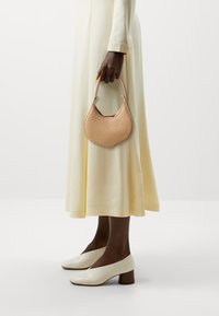 Sac à main tissé beige avec une silhouette courbée et un motif texturé, tenu dans une main. Le modèle porte une robe crème et des chaussures à talons blancs.