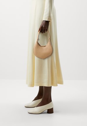 Sac à main tissé beige avec une silhouette courbée et un motif texturé, tenu dans une main. Le modèle porte une robe crème et des chaussures à talons blancs.