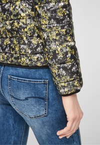 Pufjakke med et sort, gult og gråt blomsterdesign; har quiltet tekstur og ribbede manchetter. Bæres med blå denimjeans.