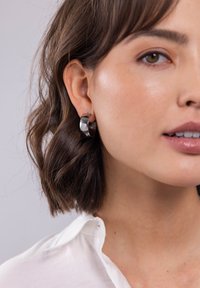 Silberner Creolenohrring mit polierter Oberfläche, der ein gedrehtes Design und eine glatte, reflektierende Fläche aufweist, getragen am linken Ohr.