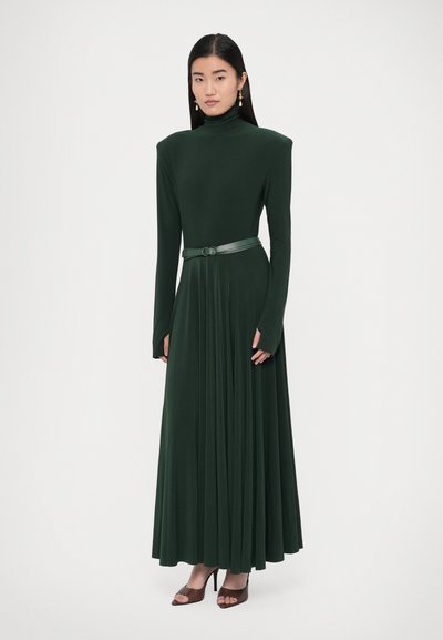 Zaļa, garām piedurknēm pieguloša kleita ar augstu apkakli, plīsētu svārku un atbilstošu ādas jostu. Modelis valkā brūnas atvērtas purngala kurpes.