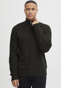 Mann mit kurzen Haaren, der einen dunkelgrünen, strukturierten, halb-zipp Pullover trägt, den Reißverschluss nahe am Hals haltend, vor einem schlichten weißen Hintergrund stehend.