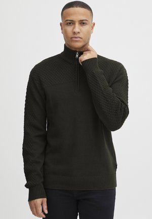 Uomo con capelli corti che indossa un maglione verde scuro a mezza zip con texture, mentre tiene la zip vicino al collo, in piedi di fronte a uno sfondo bianco uniforme.