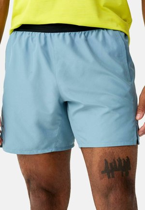 Mann trägt hellblaue Sportshorts und ein gelbes Shirt und zeigt ein schwarzes Tattoo am rechten Oberschenkel.