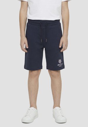 Niño con pantalones cortos azul marino con cordón y logo en la pierna izquierda, camiseta blanca y zapatillas blancas, de pie frente a un fondo liso.