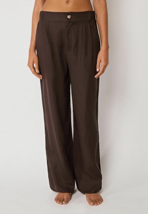 Femme portant un pantalon ample taille haute marron foncé avec un bouton et des passants pour ceinture, debout pieds nus sur un fond uni.