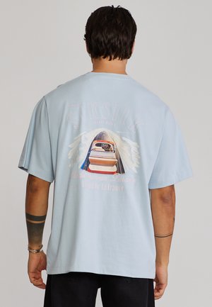 Sötét hajú férfi világoskék SikSilk pólóban, hátulnézetben, amelyen hajó grafika és szöveg látható.