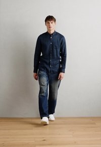 Denim shirt with button closure, dark blue, paired with faded blue jeans. Worn with white sneakers. Minimalist setting with light wood floor.