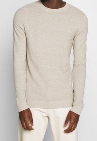 Man wearing a beige ribbed knit long-sleeve sweater and off-white pants, standing against a plain light background.