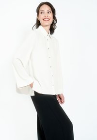 Biała koszula z długim rękawem z zapięciem na guziki, z klasycznym kołnierzykiem i dwiema bocznymi kieszeniami, w połączeniu z czarnymi spodniami o szerokich nogawkach.