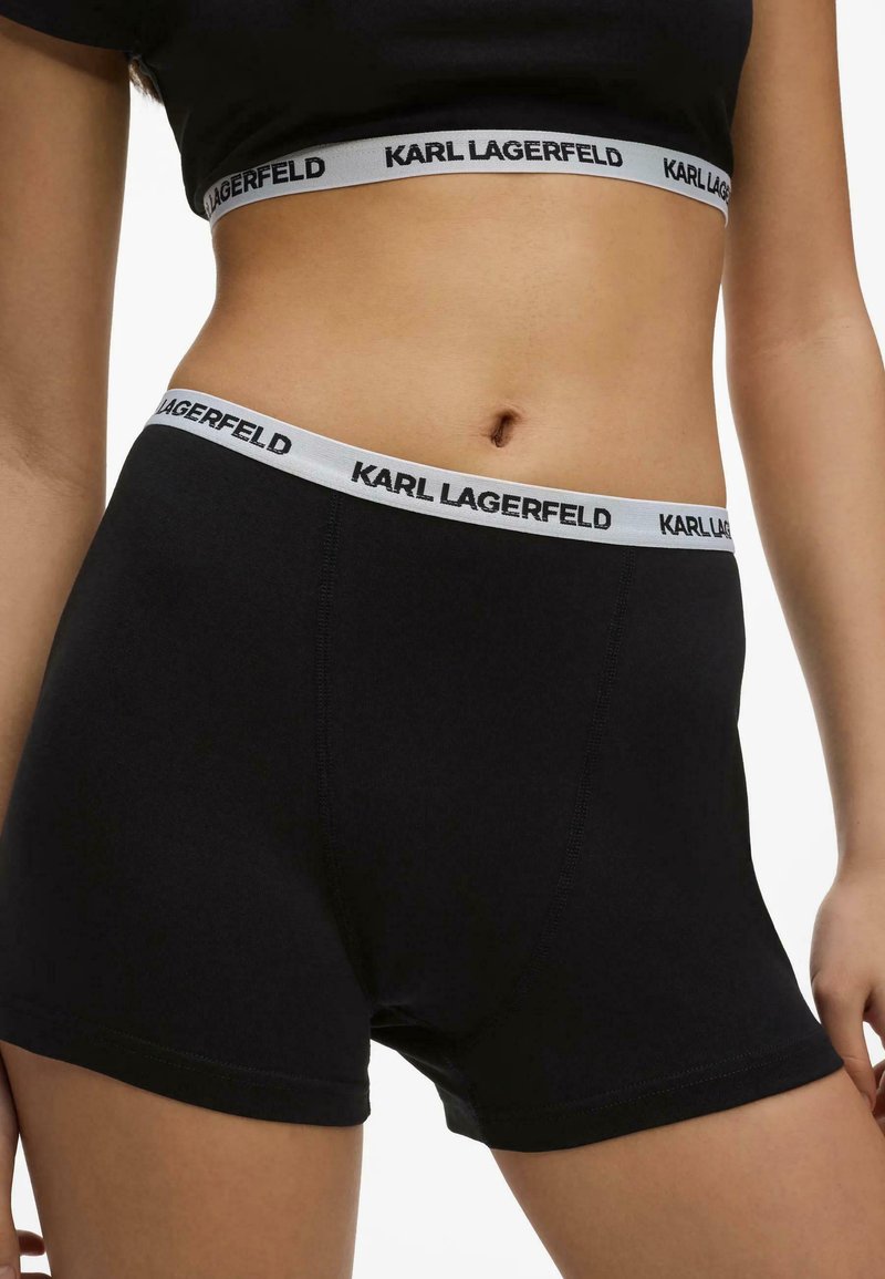 Gros plan d’une personne portant un short noir à taille élastique marqué Karl Lagerfeld et un top court assorti sur fond blanc.