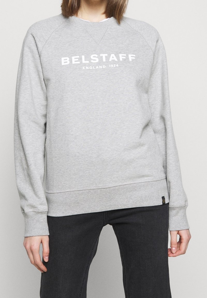 Personne portant un sweat-shirt gris clair avec le texte « BELSTAFF ENGLAND 1924 », associé à un pantalon noir, debout devant un fond uni.