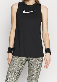 Top atletico senza maniche nero con logo Nike bianco, abbinato a leggings verdi e neri a fantasia. Presenta un tessuto morbido e una vestibilità comoda.