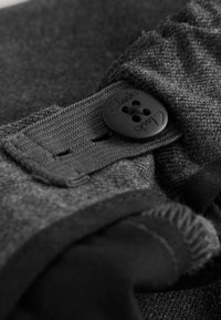 Zwarte stof met een zachte textuur, met een knop met het merk "Clarks" en een gestikte band. Close-up focust op de details van de knop en de stof.