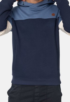 Hombre con una sudadera con capucha a bloques de color azul marino, azul claro y beige con bolsillos delanteros, ajustando la capucha sobre un fondo blanco.