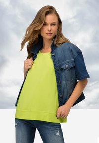 Jeansjack met korte mouwen, gecombineerd met een heldergroen topje. Model draagt slim-fit blauwe jeans. Casual, hedendaagse look.