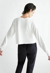 Top bianco a maniche lunghe cropped, realizzato in materiale morbido, presenta una vestibilità rilassata e polsini a coste. Indossato con pantaloni cargo neri a vita alta.