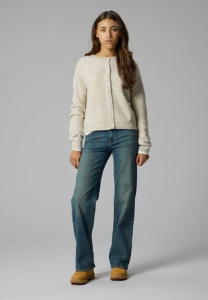 Jonge vrouw staand, gekleed in een beige button-down vest, blauwe spijkerbroek en bruine laarzen tegen een effen grijze achtergrond.