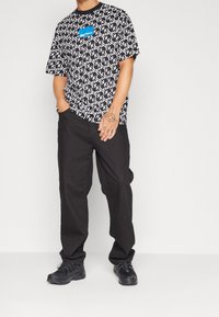 Svartvitt grafiskt t-shirt med ett upprepande mönster, kombinerat med svarta byxor och svarta sneakers. Iögonfallande blå logotyp på t-shirten.