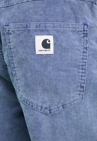 Tarfă albastră de corduroy cu dungi verticale, având o etichetă albă pătrată cu un logo negru pe buzunarul din spate. Buzunarul este de formă rectangulară.