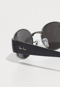 Pyöreät mustat aurinkolasit tummilla linsseillä, hopeiset kehysyksiköt ja näkyvä Ray-Ban-logo paksussa mustassa sangassa.