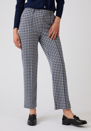 Donna che indossa pantaloni a gamba dritta a quadri blu navy e bianchi, mocassini neri e una maglia blu navy, in piedi davanti a uno sfondo chiaro.