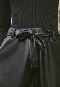 Pantaloni neri di pelle con un dettaglio della cintura annodata, dotati di una superficie liscia e di un top nero a coste aderente sopra.