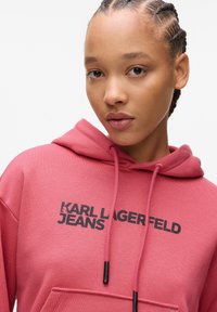 Rožinė gobtuvas, pagamintas iš minkštos medžiagos, su priekinėmis kišenėmis, raišteliais ir spausdintu juodu tekstu "KARL LAGERFELD JEANS."