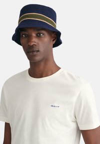 Marinevævet bucket hat med to vandrette striber i grøn og beige; ledsaget af en cremefarvet t-shirt med et lille blåt logo.