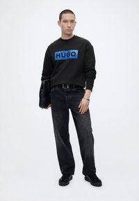 Jeune homme portant un sweat-shirt noir avec le logo bleu "HUGO", jean noir, chaussures noires, tenant un sac noir, debout devant un fond blanc.