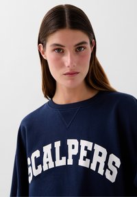 Marineblå sweatshirt med hvid "SCALPERS" skrift. Har en rund halsudskæring og ribbet kant. Blødt stof med en glat tekstur.