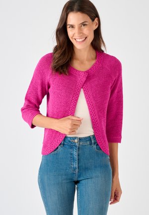 Vrouw die lacht, draag een felroze gebreid vest met driekwart mouwen over een wit shirt en een blauwe spijkerbroek, tegen een effen lichte achtergrond.