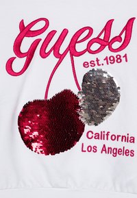 Vit sweatshirt med stort broderat "Guess"-logotyp i rosa och en paljettdesign av ett körsbär i röd och silver, med texten "California Los Angeles" nedan.
