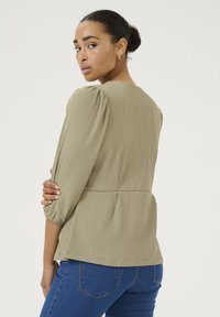 Blusa verde oliva con texture, maniche a sbuffo, vita arricciata e bordo posteriore curvato, abbinata a jeans blu aderenti.