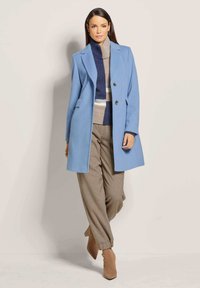 Hellblauer Wollmantel mit taillierter Passform, zwei Knöpfen und Seitentaschen, kombiniert mit einem gestreiften Pullover in Beige und Marineblau sowie beigen Hosen.