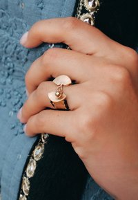 Bague de couleur or présentant un design en forme d'éventail avec des crêtes et un petit accent en forme de boule. Une main tient un tissu texturé et sombre.